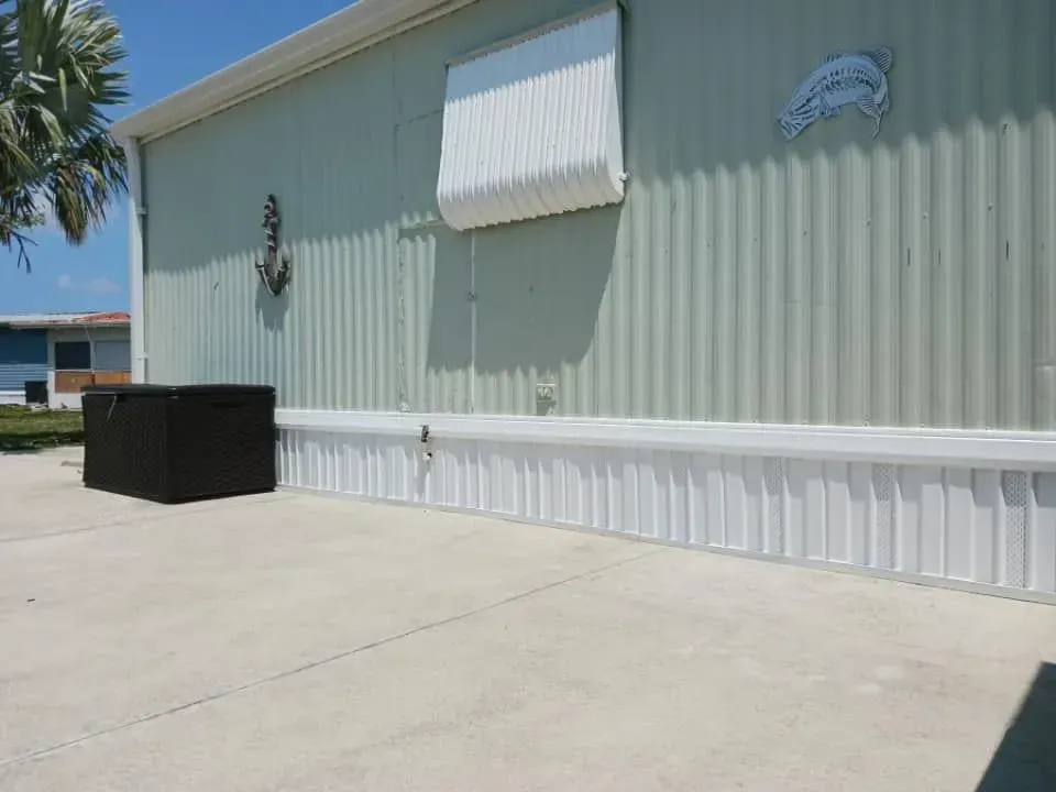 Side view of a light green mobile home with white accents, nautical decor, and a black storage chest on a concrete patio. Side view of a light green mobile home with white accents, nautical decor, and a black storage chest on a concrete patio.