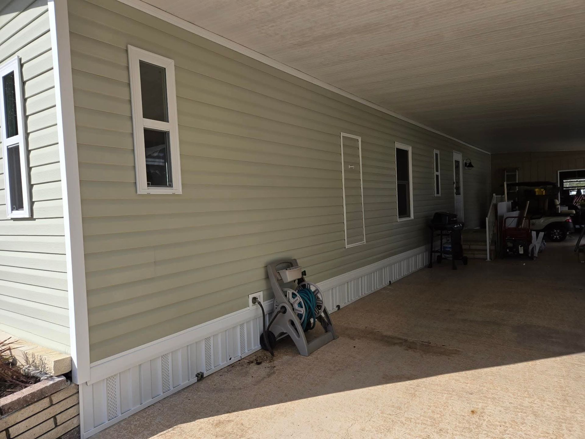 Beige mobile home exterior with white trim, carport. A gray step stool leans against the wall.
