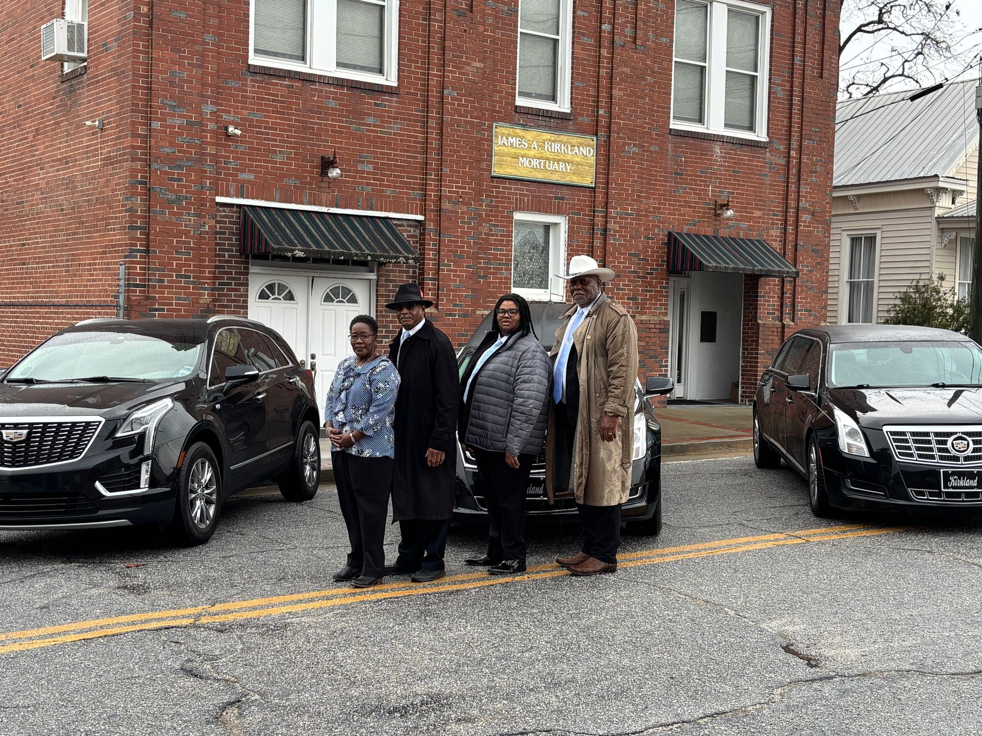 Three black hearses and three men in suits parked in front of a brick building.