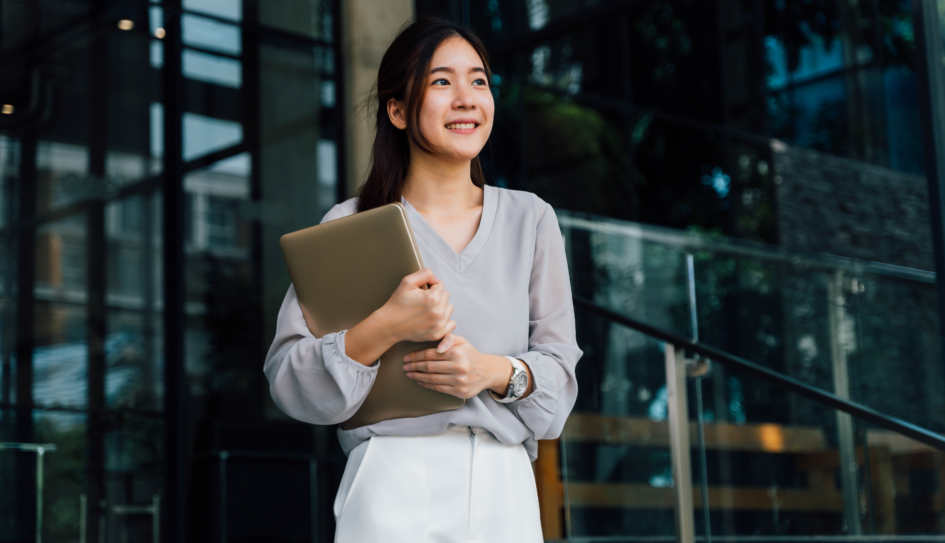 Woman with laptop smiling outside modern building.