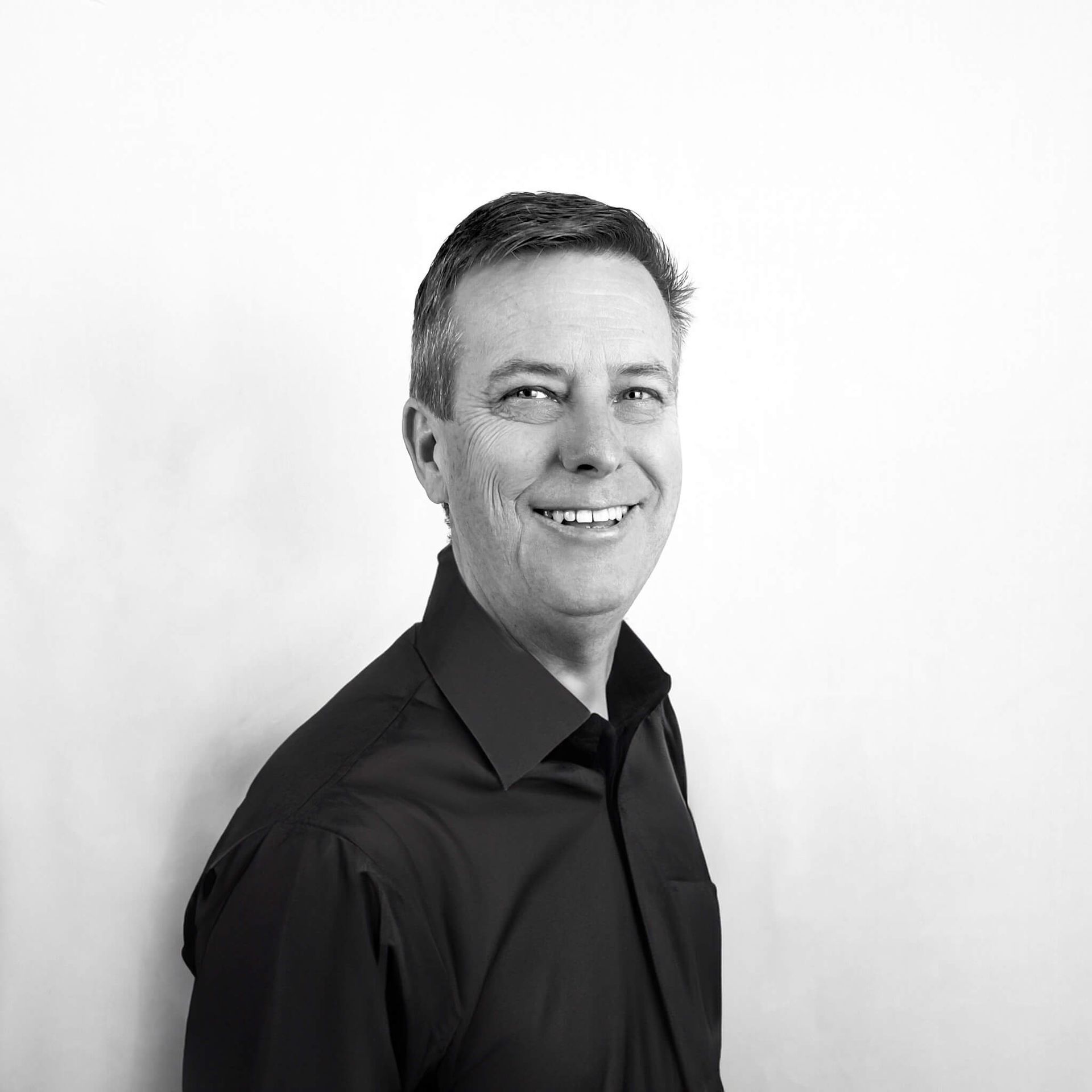 A man in a black shirt and tie is smiling in a black and white photo.