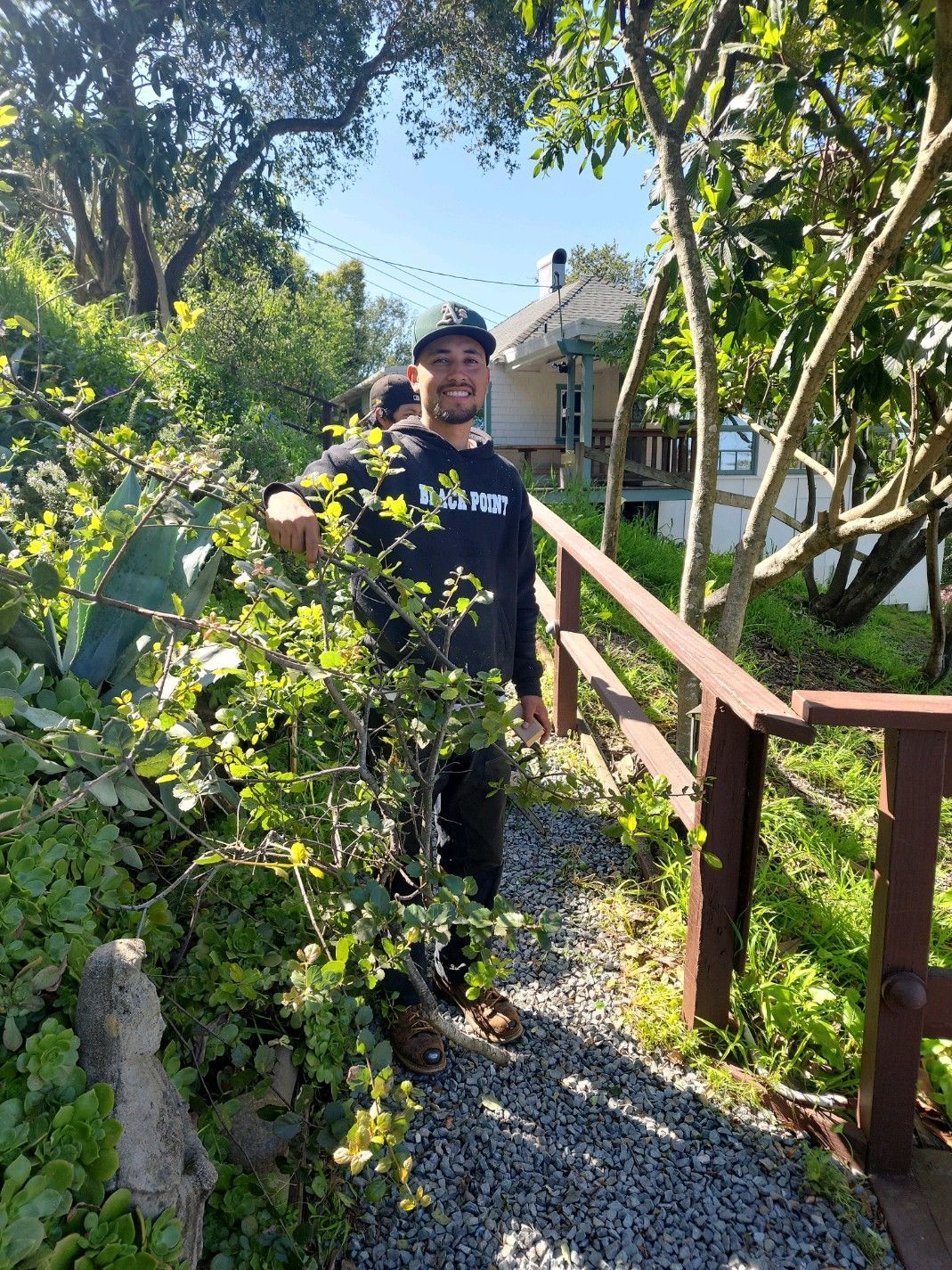 Man in black sweatshirt stands on gravel path in a sunny, green garden, leaning on a wooden railing.