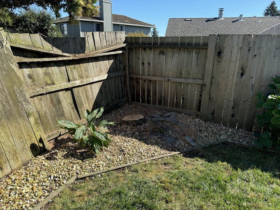 A corner of a backyard with a damaged wooden fence, gravel, and grass.