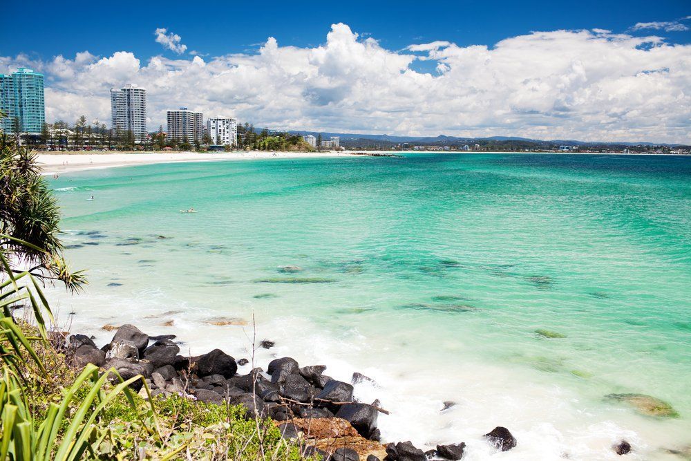 Turquoise ocean meets sandy beach under a bright blue sky with clouds, buildings on the horizon.