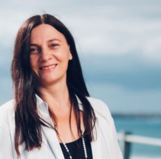 Woman with long dark hair smiles outside, wearing white shirt and necklace; ocean background.