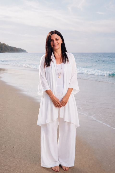Woman with long dark hair smiles outside, wearing white shirt and necklace; ocean background.