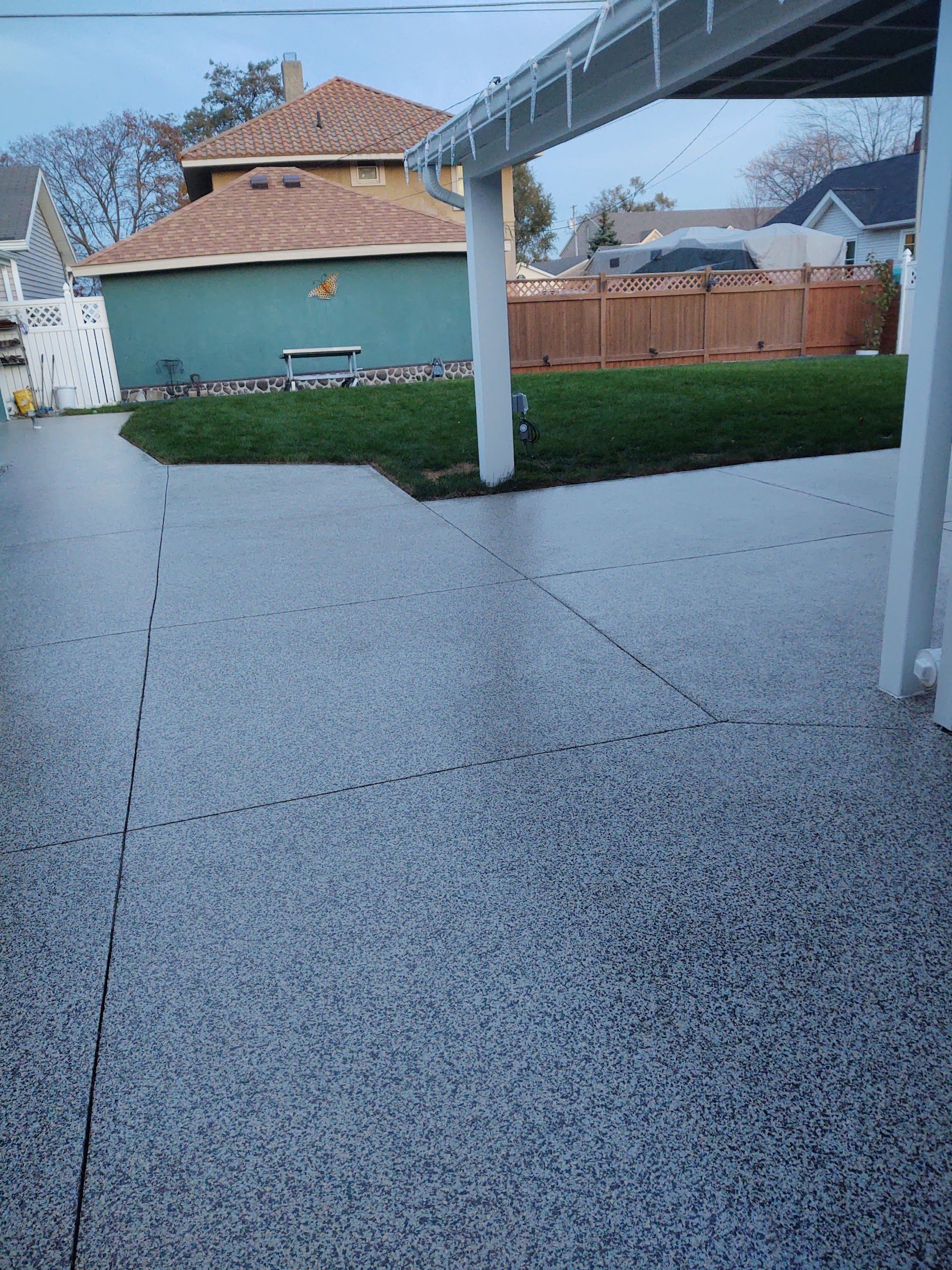 A patio with gray speckled flooring, bordered by a green lawn, wooden fence, and a house with a tan roof at dusk.