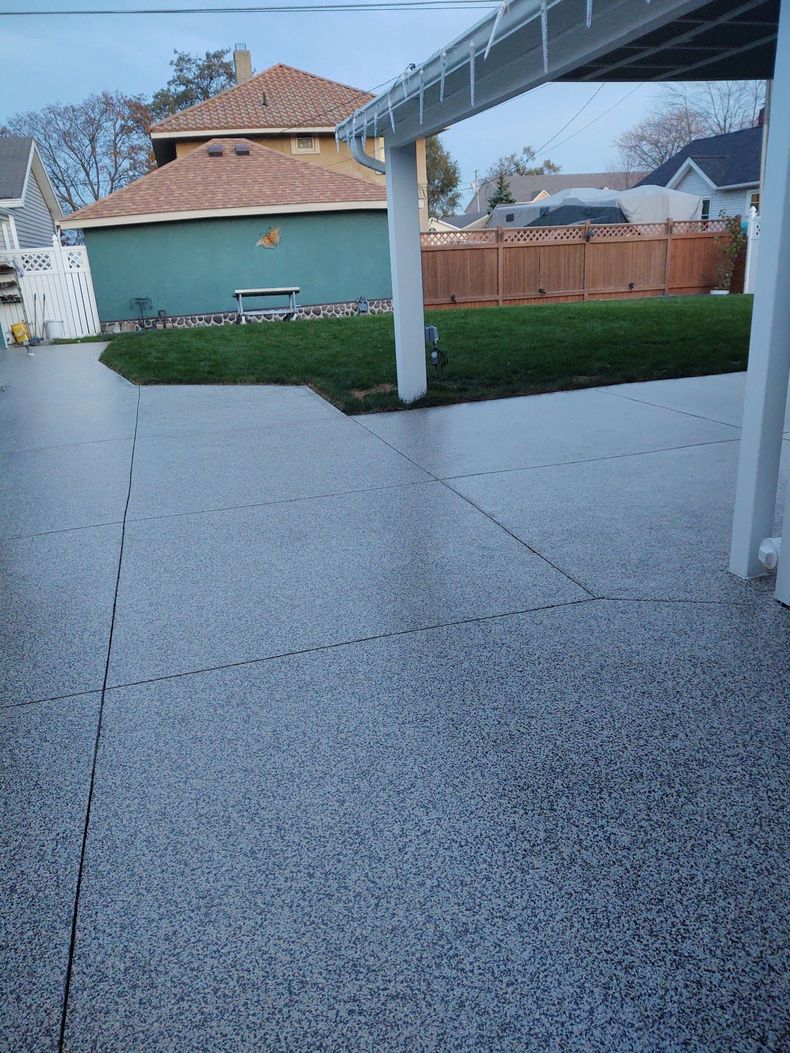 A patio with gray speckled flooring, bordered by a green lawn, wooden fence, and a house with a tan roof at dusk.