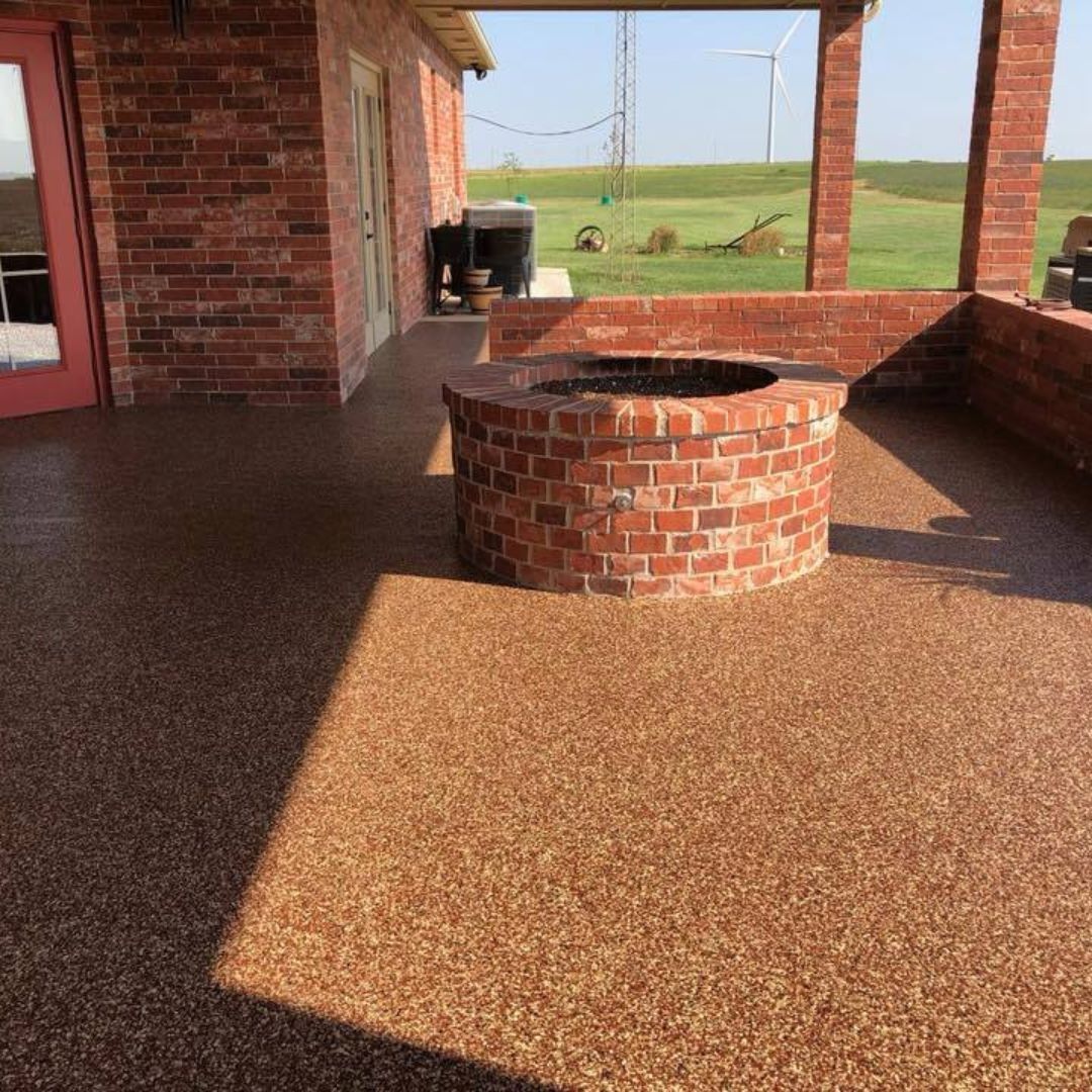 A covered brick patio with a round brick fire pit and speckled brown flooring, overlooking a grassy field.