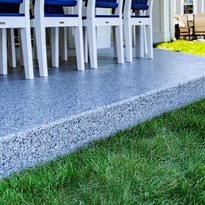 White chairs with blue cushions sit on a grey speckled patio overlooking a green lawn.