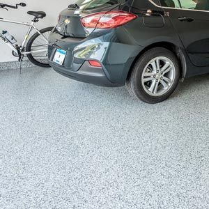 A dark gray car parked in a garage with a light gray speckled epoxy floor and a bicycle leaning against the wall.
