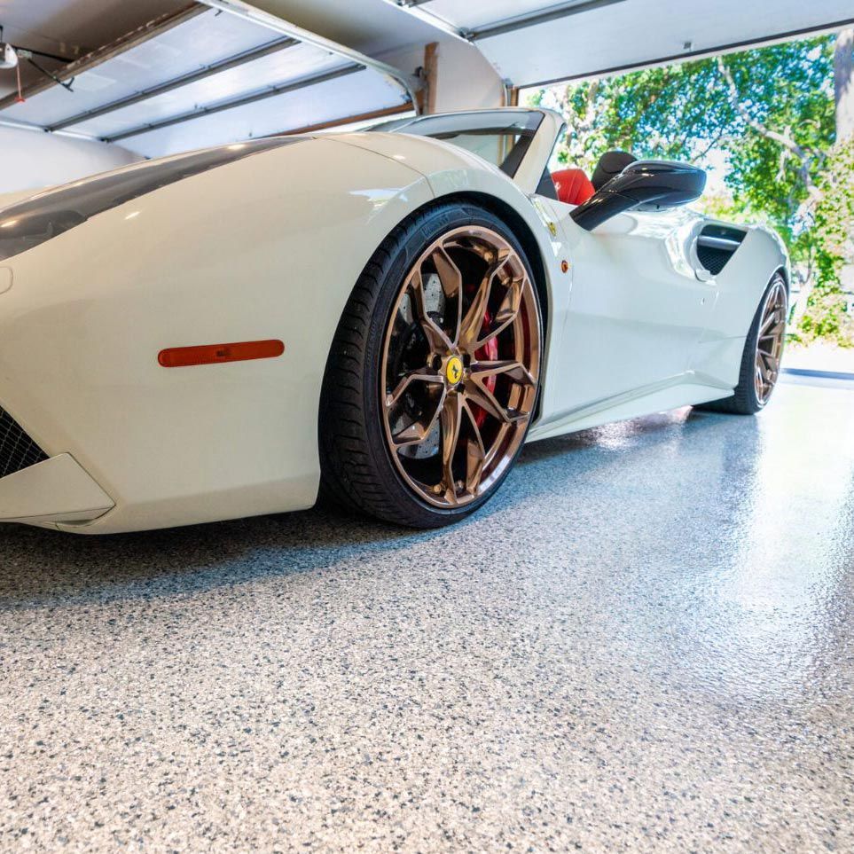 A cream-colored sports car with bronze wheels parked inside a residential garage with a speckled epoxy floor.