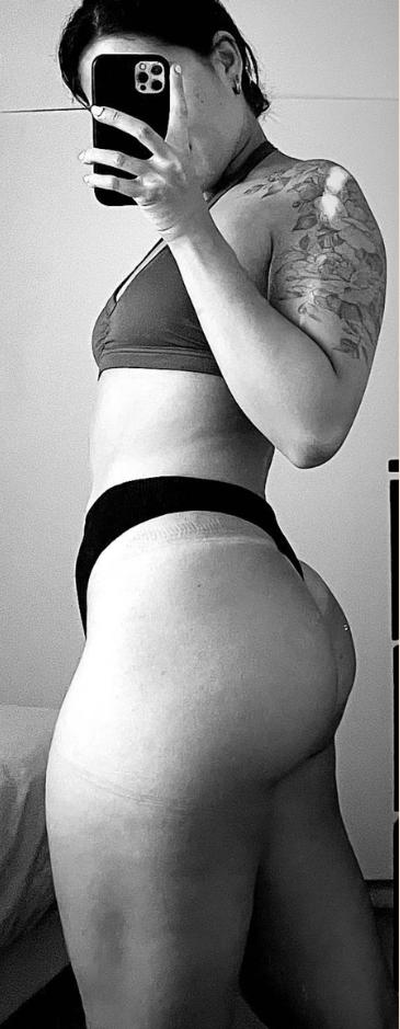 Woman in a bra and underwear, taking a mirror selfie. Black and white photo, tattoo on arm.