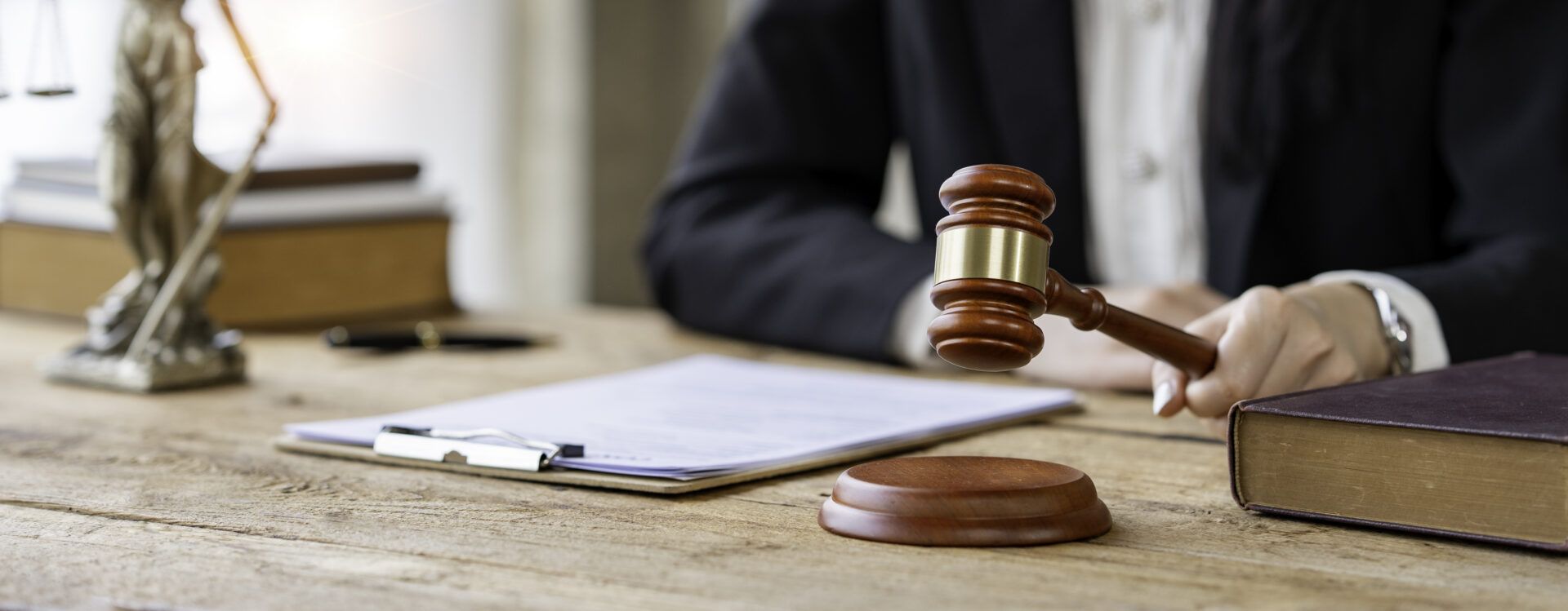 A person in a suit holding a gavel on a wooden desk with a clipboard and law book.