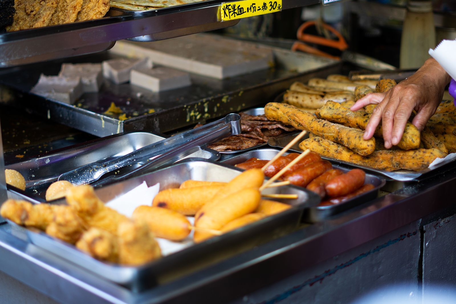 Puesto de comida con diversos productos fritos, como salchichas, perritos calientes y pasteles rectangulares. Una mano alcanza la comida.