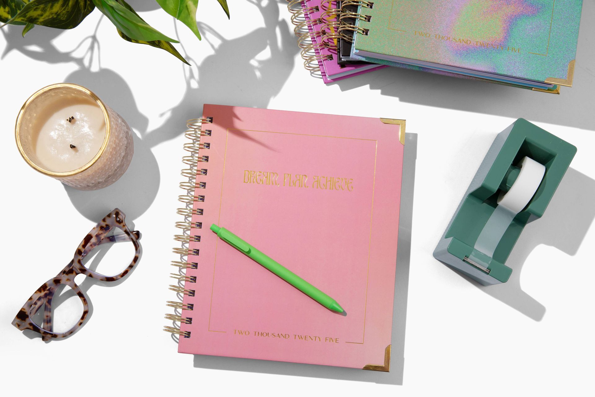 A pink notebook with a green pen and glasses on a table.