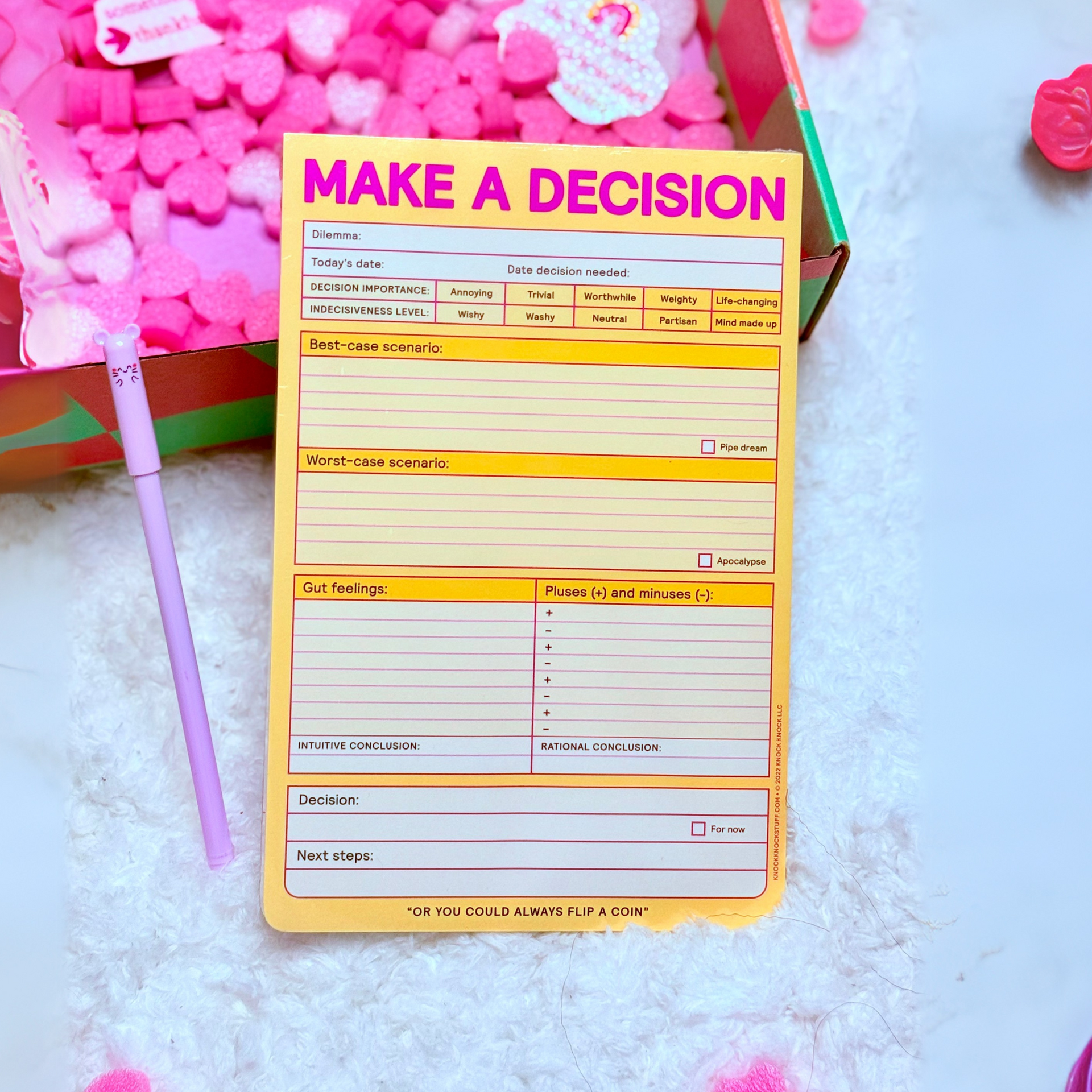 A notepad that says `` make a decision '' is sitting on a table next to a box of pink hearts.
