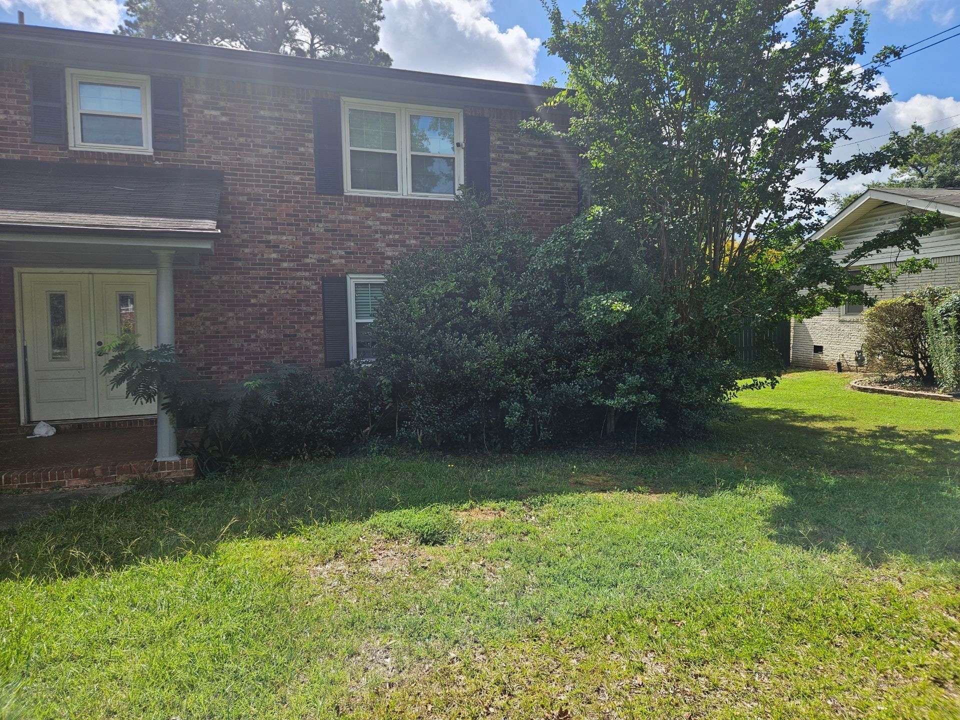 Brick building with green grass, bushes, and a partial view of a neighboring white house under a partly cloudy sky.