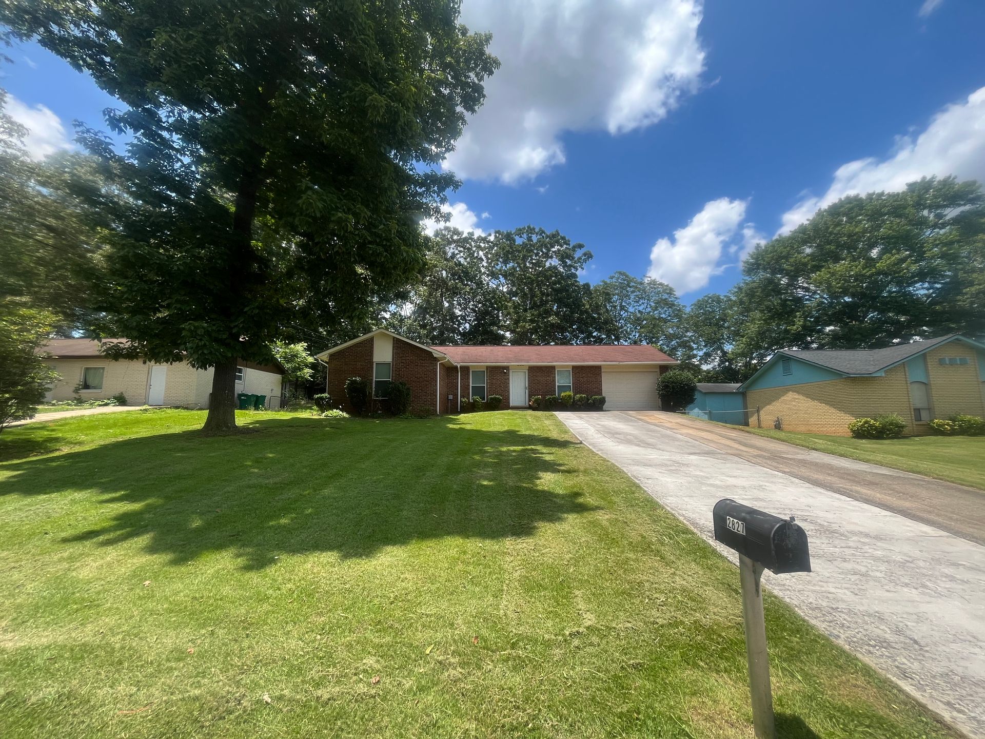 A one-story brick house with a driveway and green lawn under a partly cloudy sky.