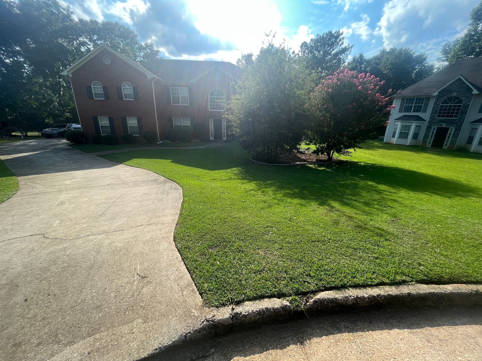 Two-story brick house with green lawn, driveway, and trees under a sunny sky.
