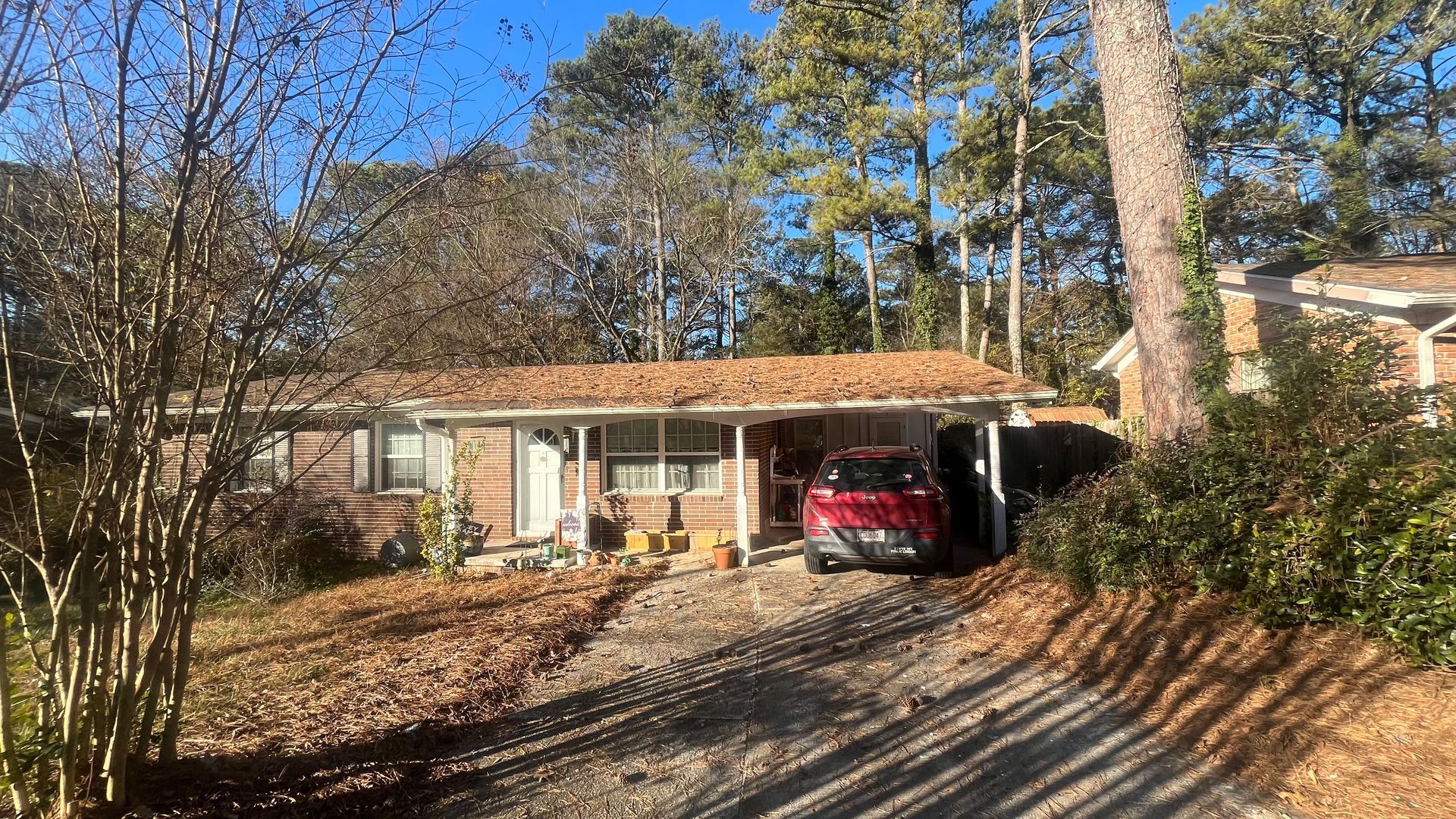 Brown one-story house with a carport; red car parked under it. Driveway, trees, and blue sky visible.