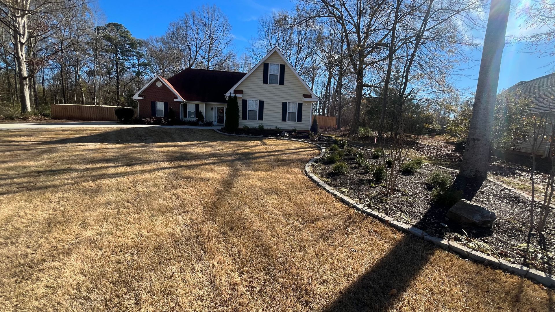 House with a red roof, tan siding, and black shutters, set in a yard with brown grass and trees under a blue sky.