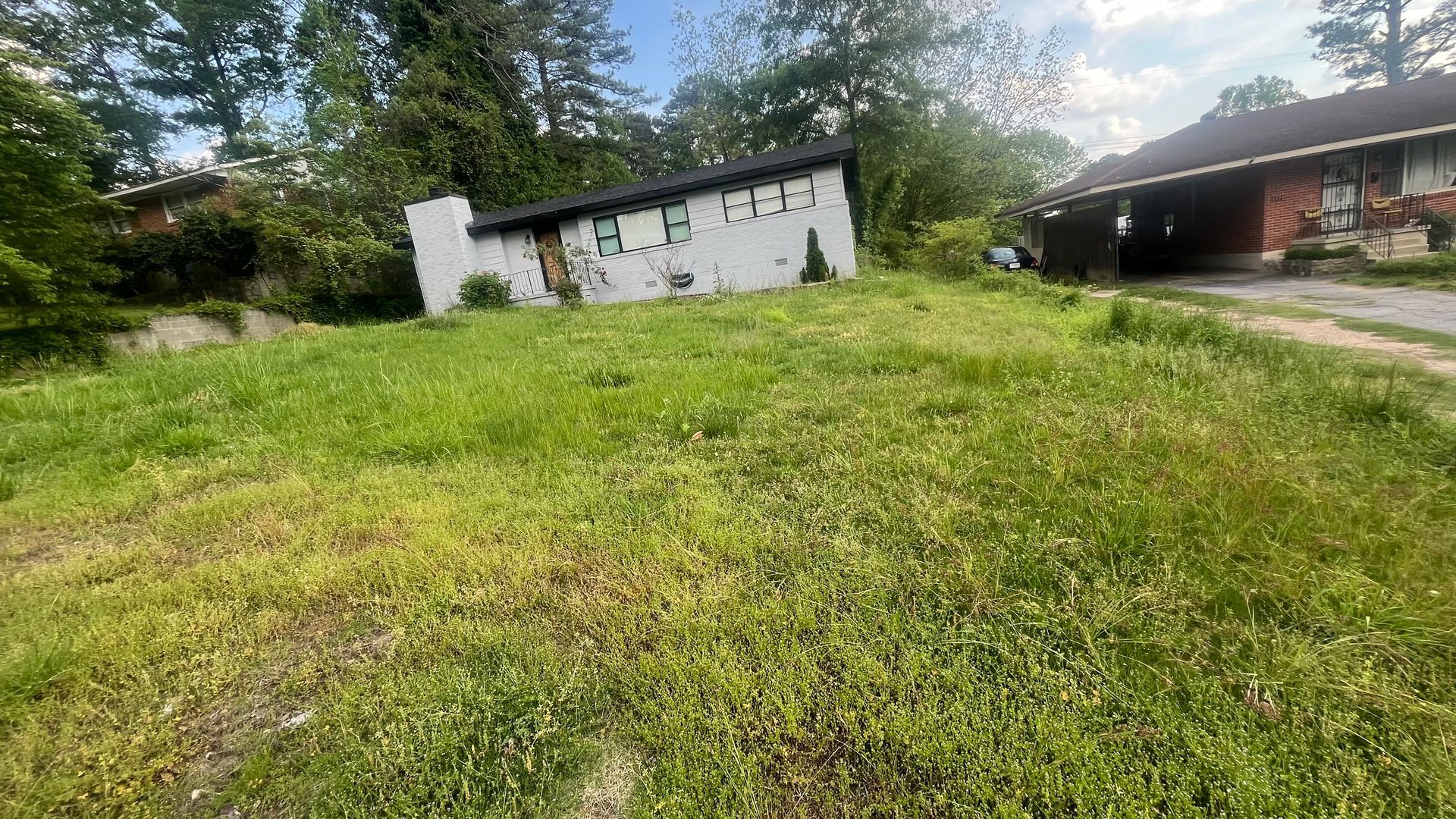 Overgrown grassy yard with a dilapidated white building, trees, and a brown house.