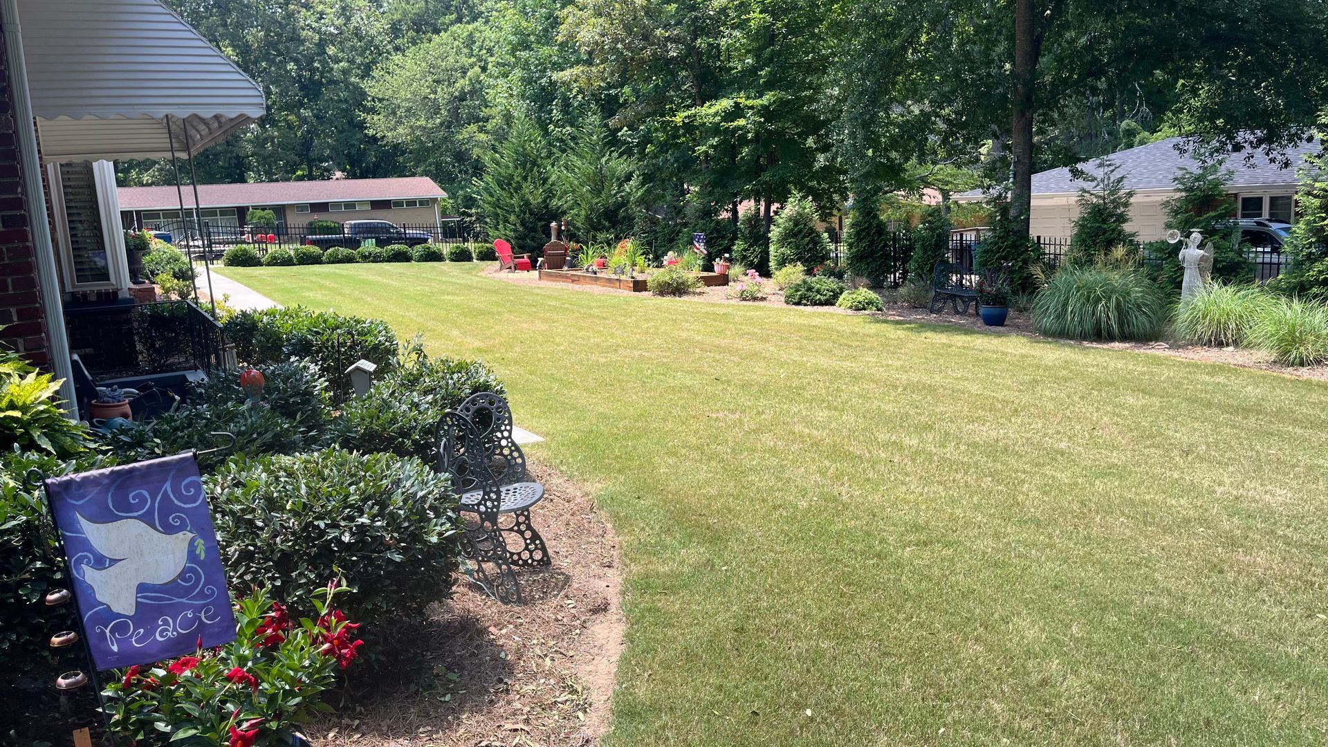 Lush green lawn with garden beds, a flag with a dove, and trees in a residential neighborhood on a sunny day.