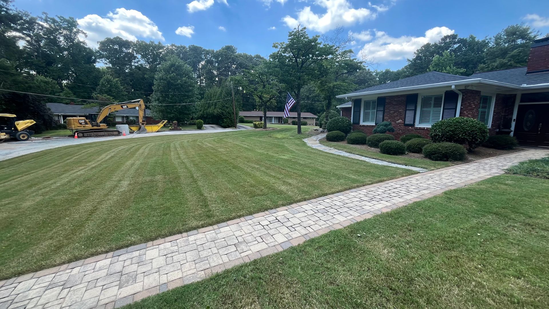 A brick house with a paved walkway, green lawn, and construction equipment in the background under a blue sky.