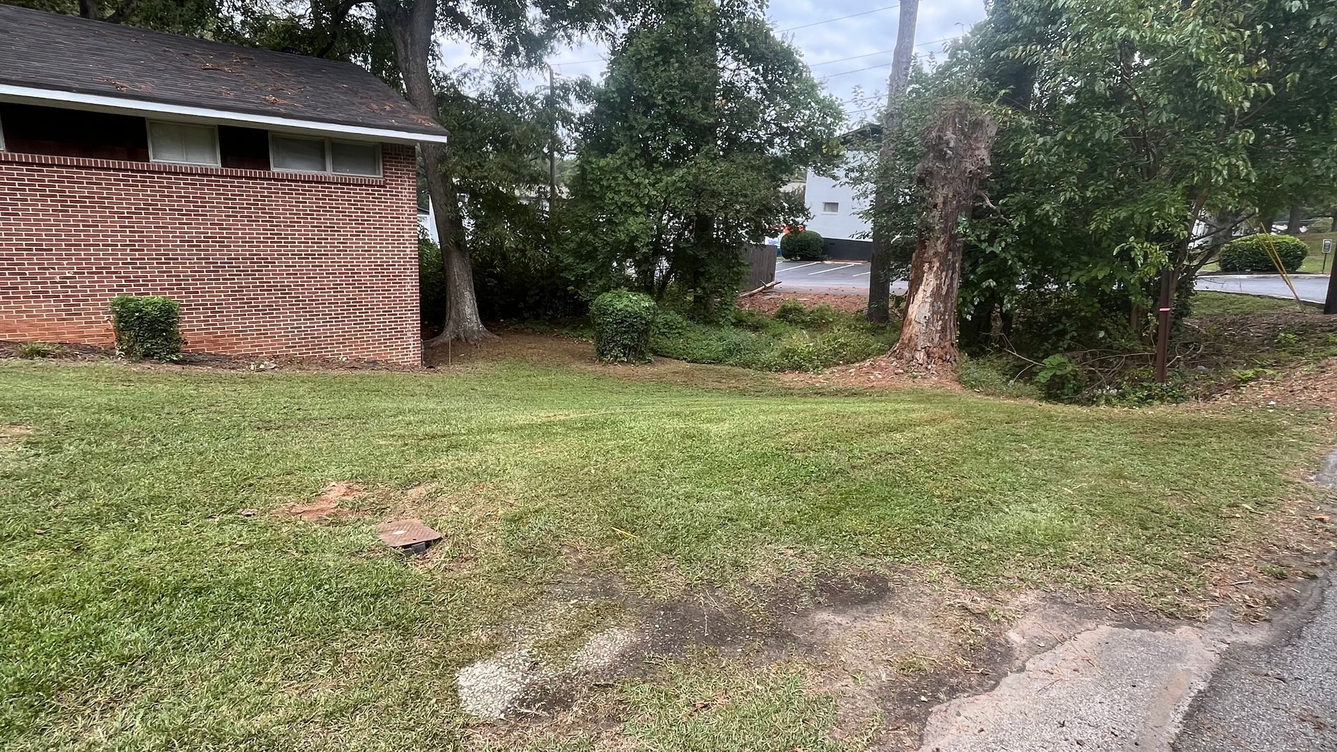 Lawn with brick building on left, trees in background and a patch of discolored soil and asphalt in the foreground.