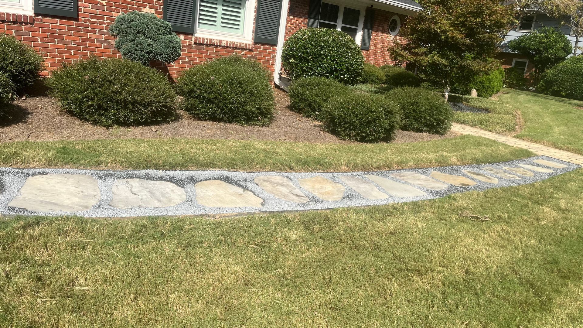 Stone pathway curves through a green lawn, bordering a brick house and trimmed shrubs.