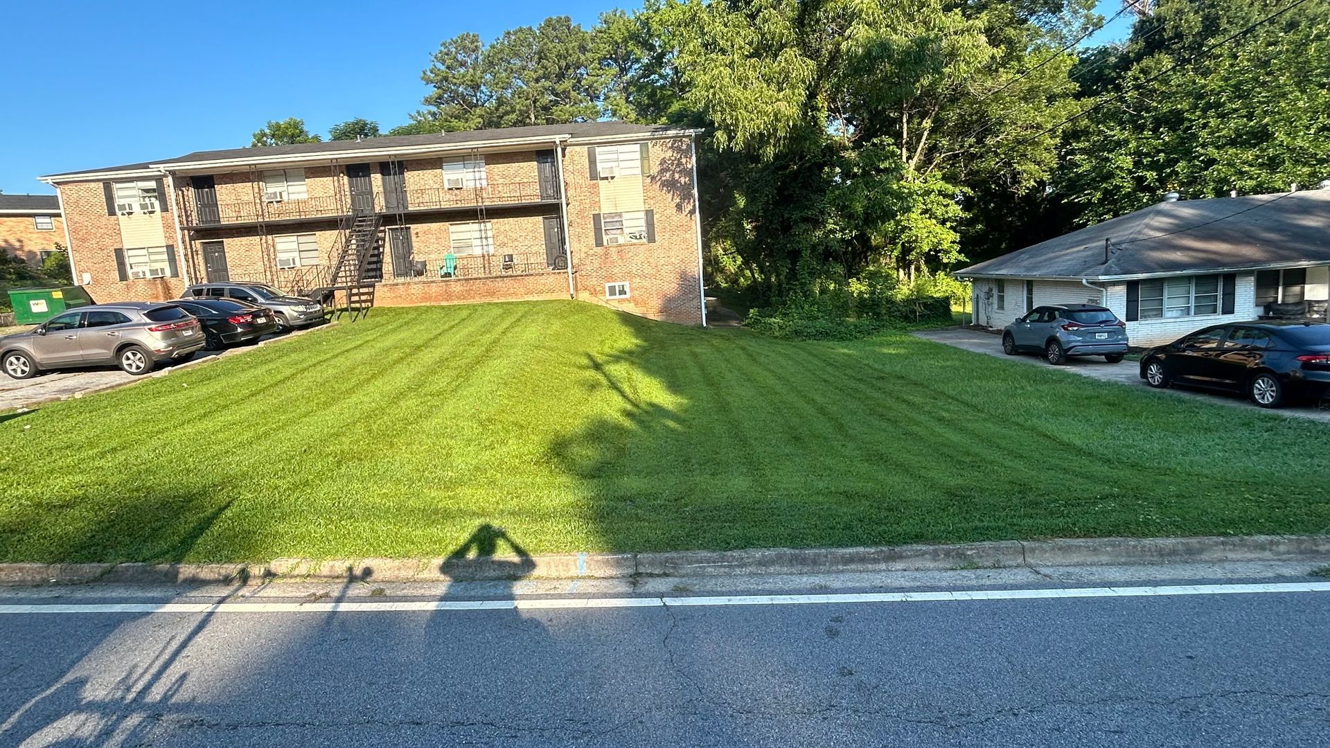 An apartment building next to a single-story house with a freshly mowed lawn, cars parked on either side, sunny day.