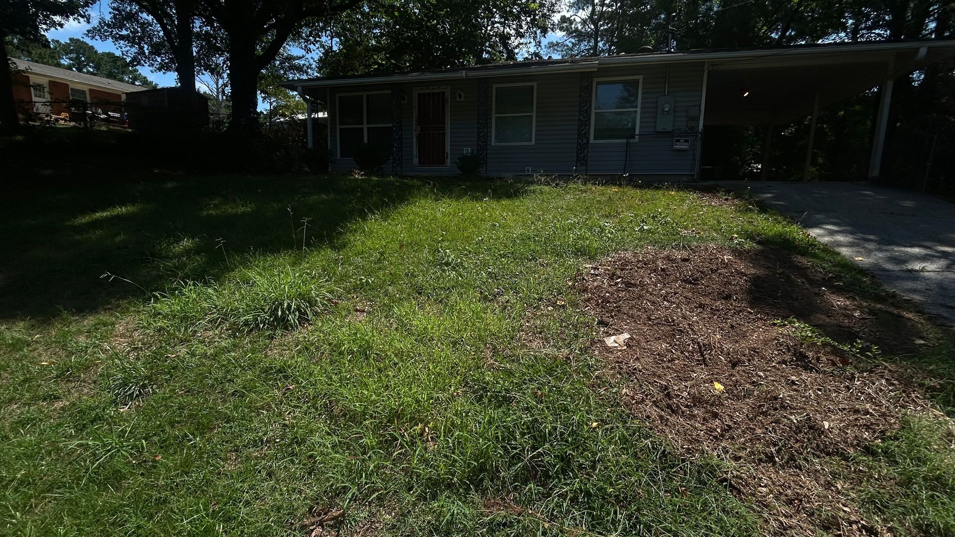 A single-story house with a weedy front lawn and a driveway under a carport.