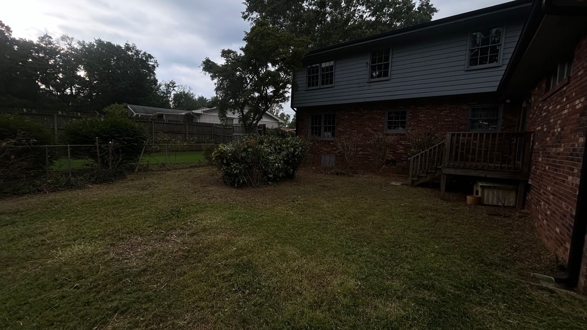 Backyard with brick and gray shingled house, grassy lawn, bushes, trees, and cloudy sky.