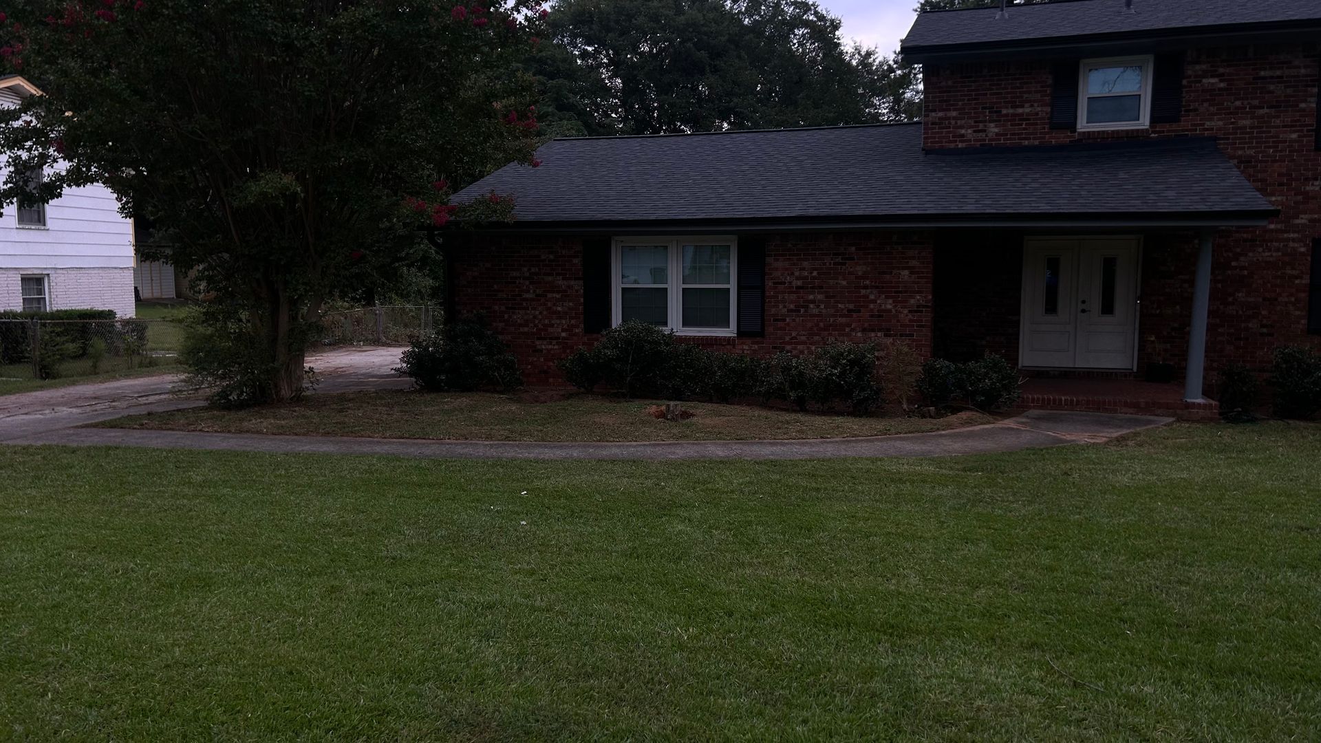 Brick two-story house with green lawn, trees, and cloudy sky. Driveway leads to a second, smaller building.