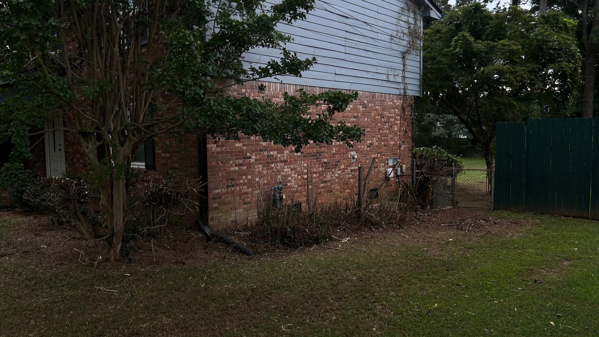 Brick and white building in a grassy yard, with a green fence and trees.