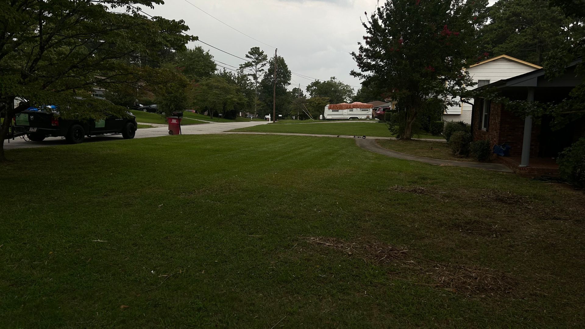 Grassy suburban yard with a road, trees, houses, and a cloudy sky.
