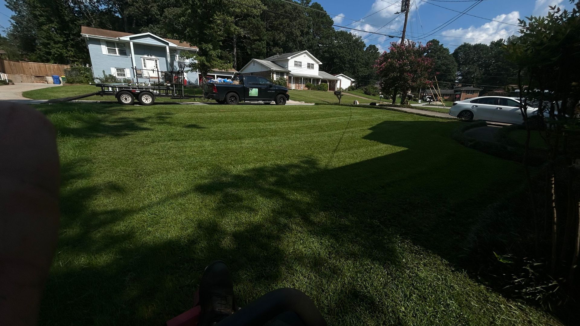 Lush green lawn with a few cars and houses in the background on a sunny day.