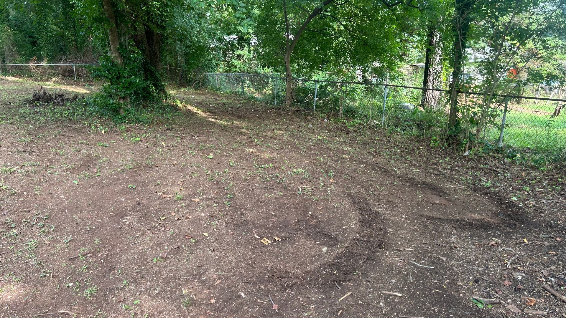 Dirt path leading through a wooded area; trees and a chain-link fence visible.