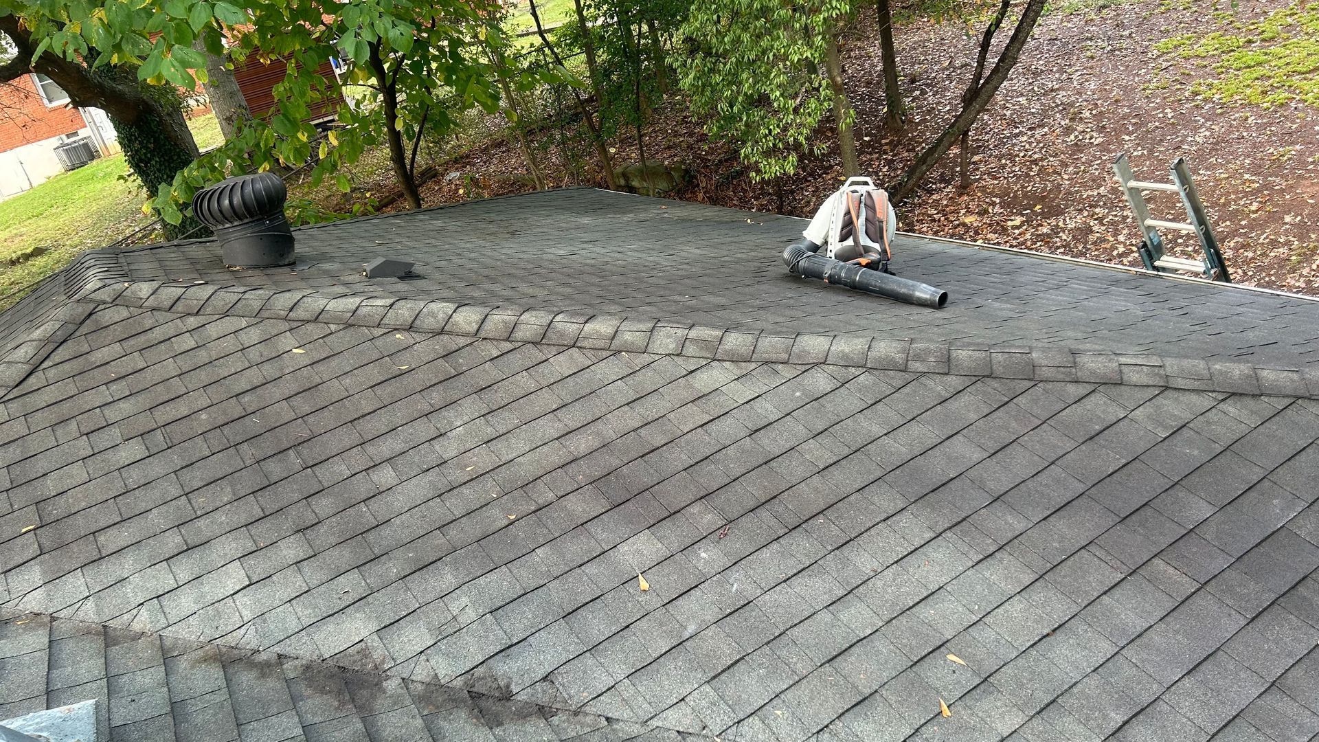 Roofer using a leaf blower on a gray shingle roof, with trees in the background.