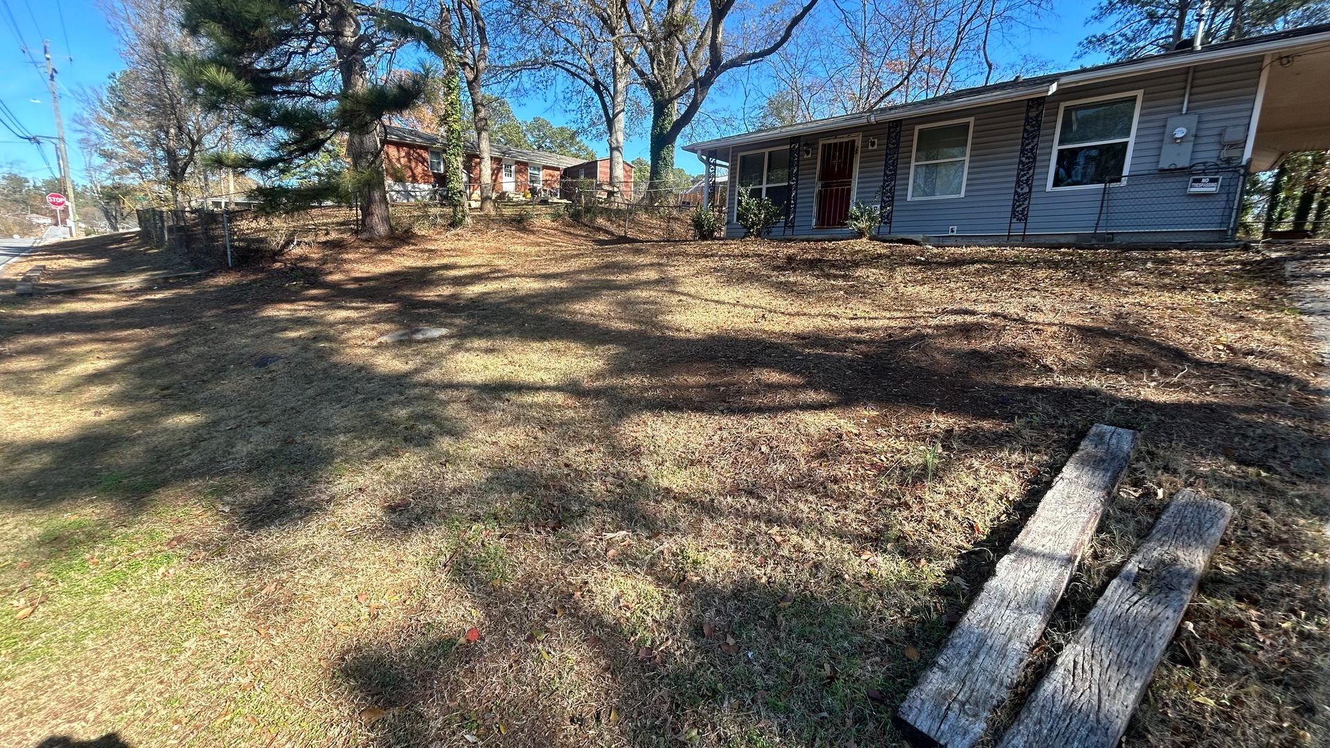 Grassy yard in front of a blue house with trees, with two wooden planks in the foreground.