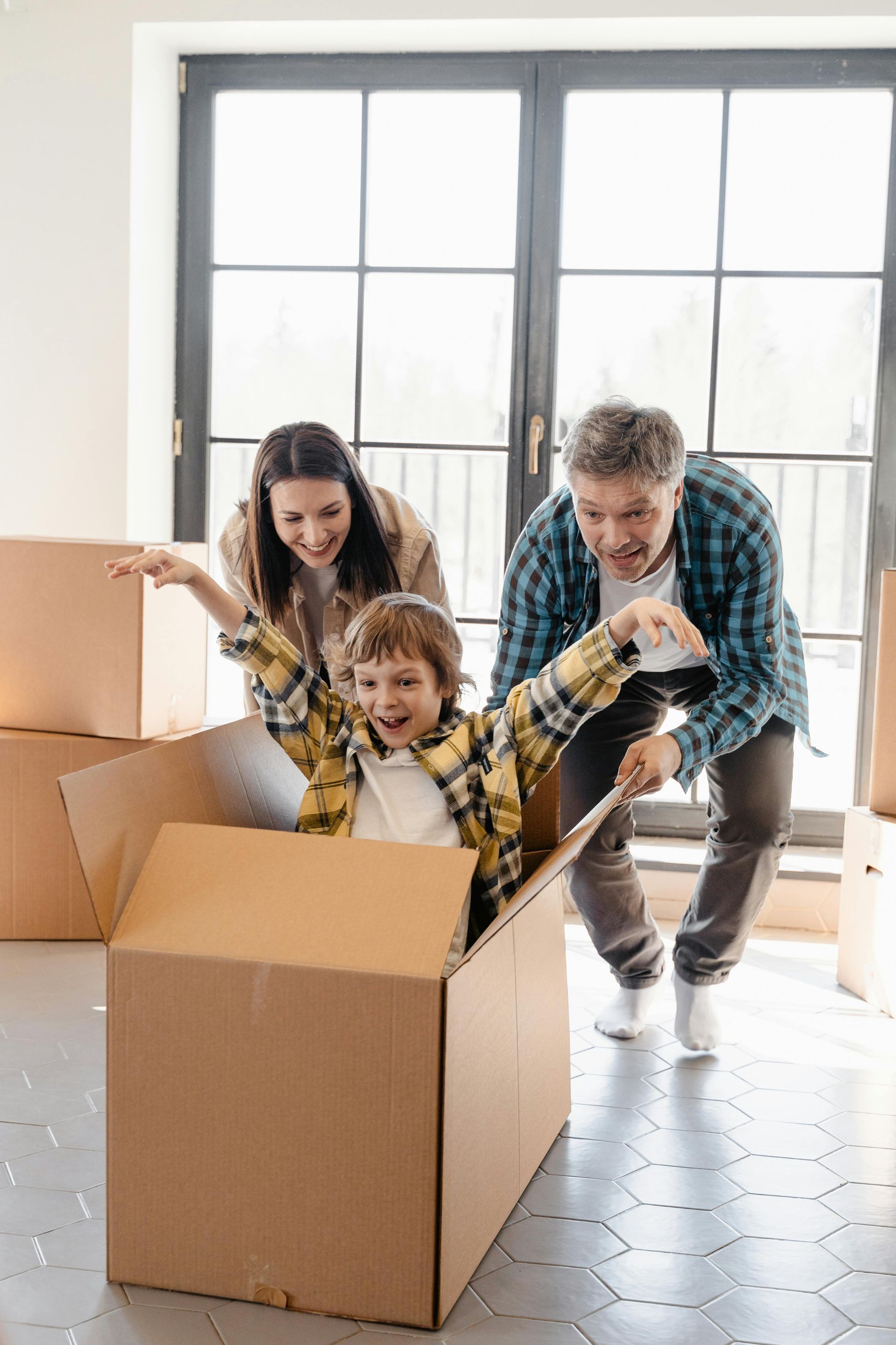 Family plays with child in a cardboard box, smiling excitedly, by a window in a room with moving boxes.