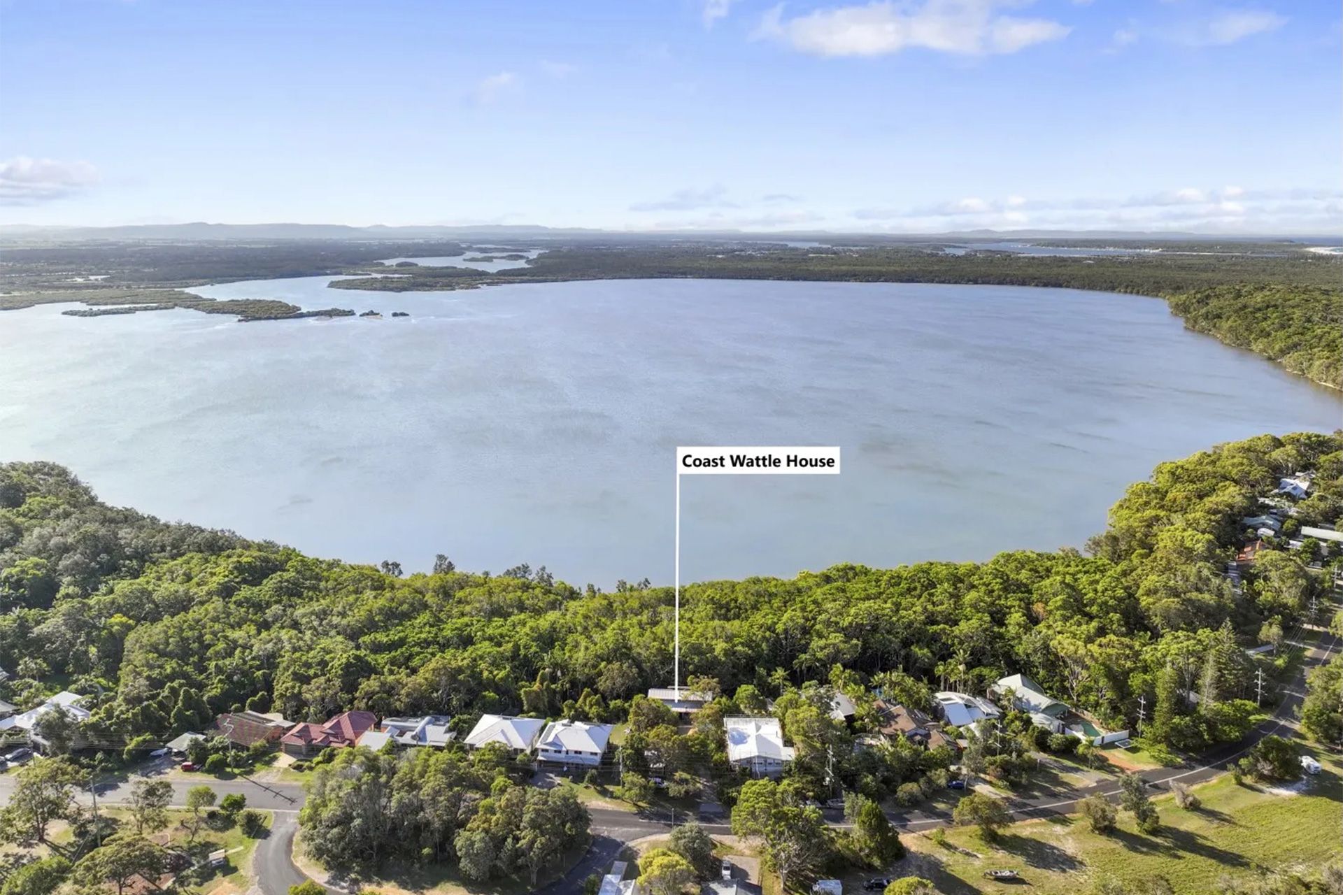 An aerial view of homes near a large lake, surrounded by trees under a bright blue sky. The text says "Coast Wattle House".
