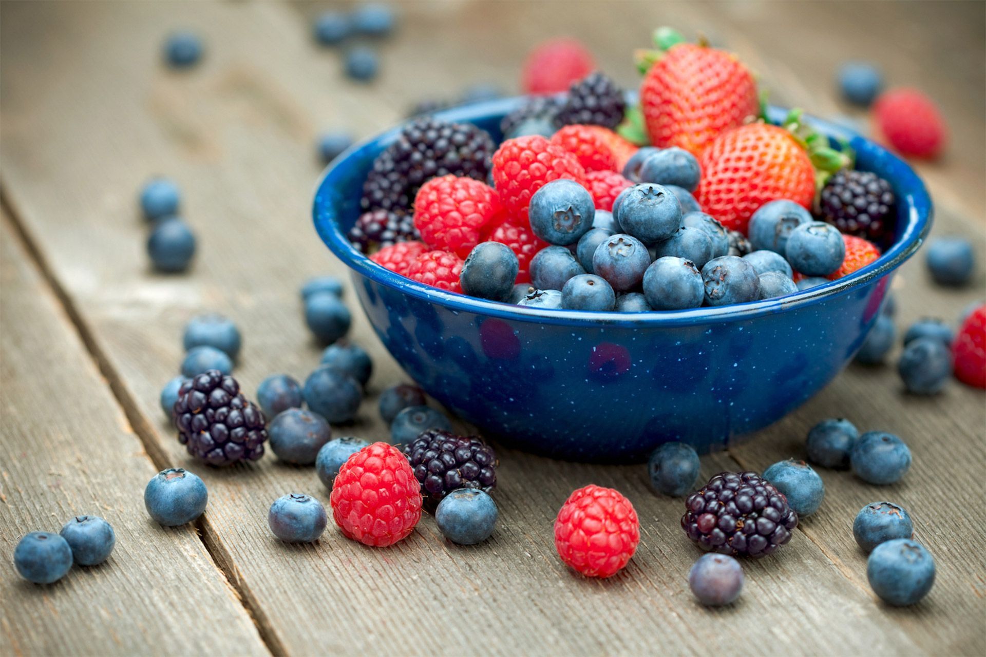 Blue bowl overflowing with blueberries, strawberries, raspberries, and blackberries, scattered on a rustic wooden surface.