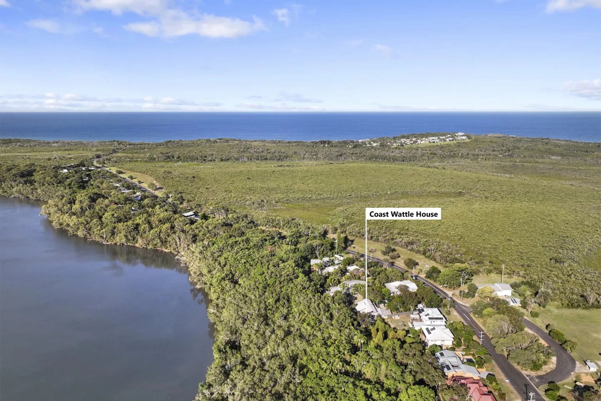 Aerial view of a coastal landscape. A lake is on the left, with houses and a road in the foreground. The ocean is in the background.