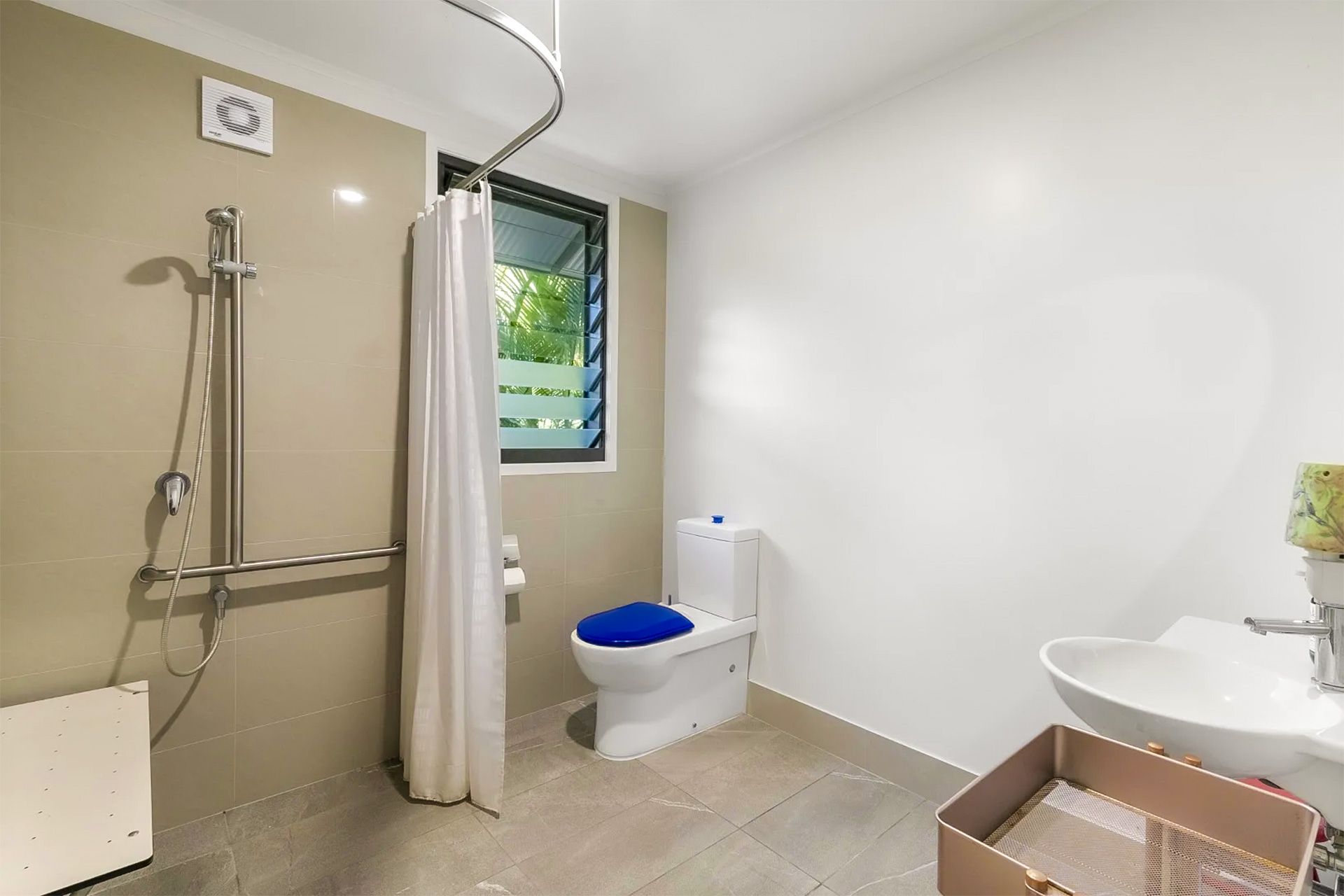 Accessible bathroom with shower, toilet, and sink. Beige walls, light gray floor tiles, and a partially open window.