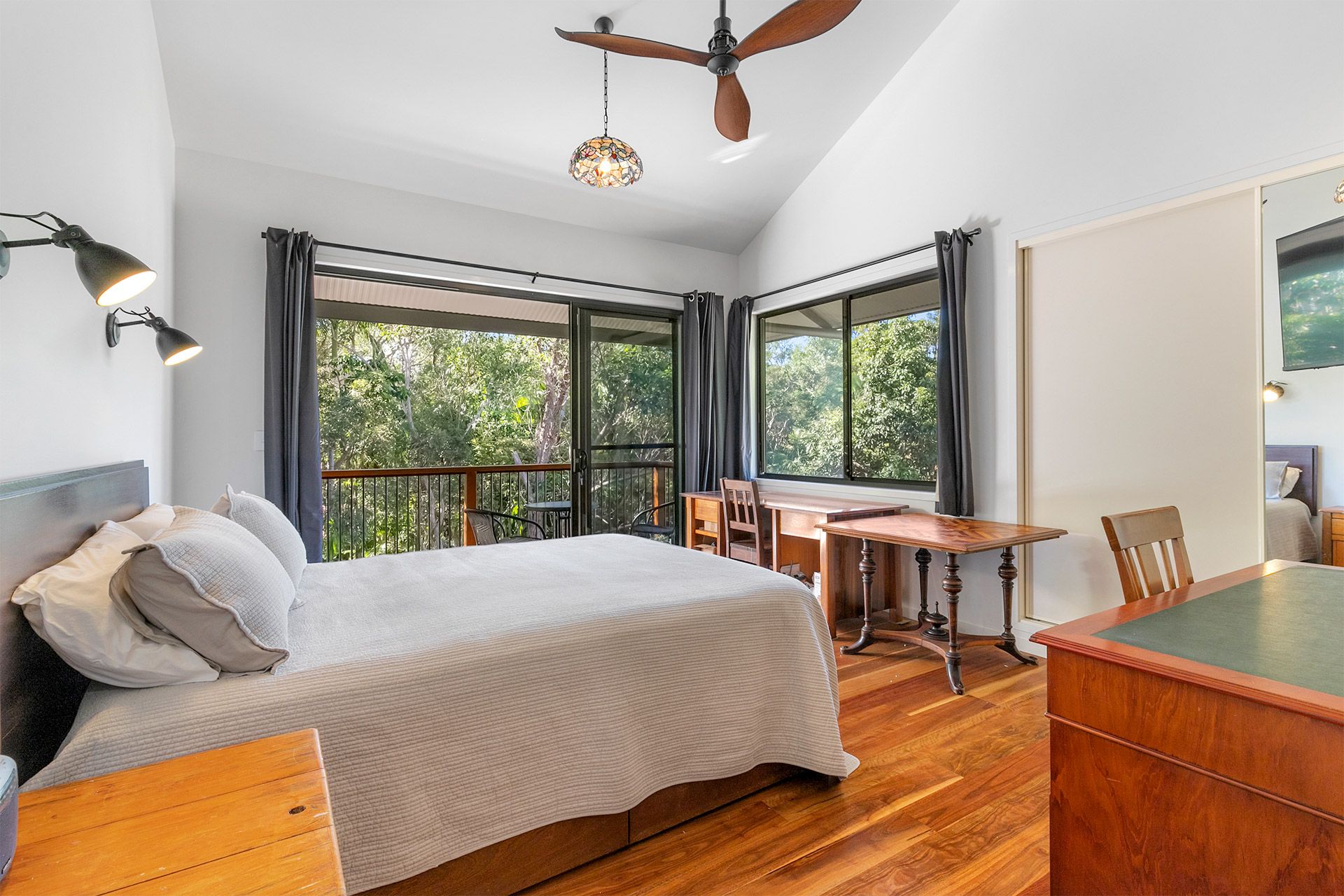 Bedroom with a bed, wooden desk, large windows overlooking trees, and a ceiling fan. Sunlight streams in.