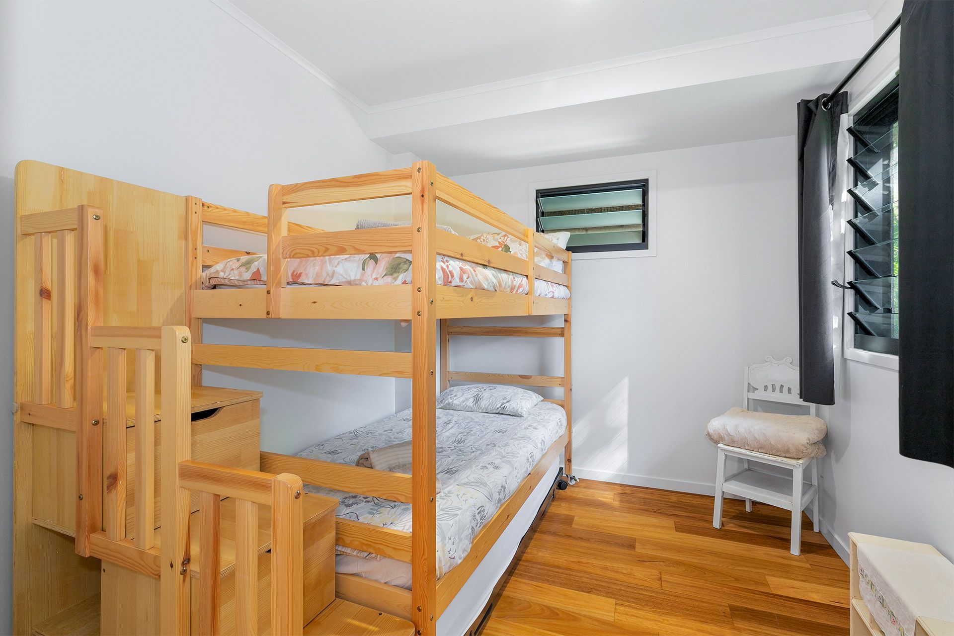 A bedroom with wooden bunk beds, wooden floors, and a white chair. There's a window with dark curtains.