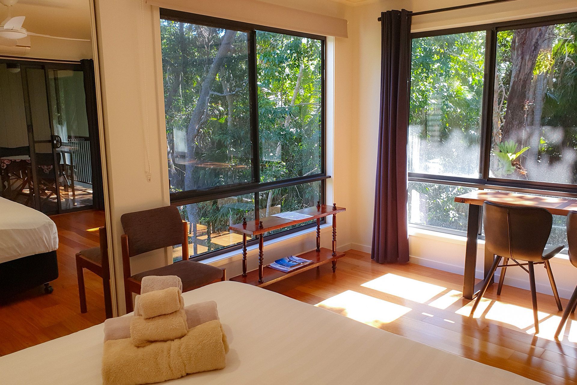 Bedroom with large windows overlooking trees. A bed is in the foreground with folded towels, chairs, and a small table.