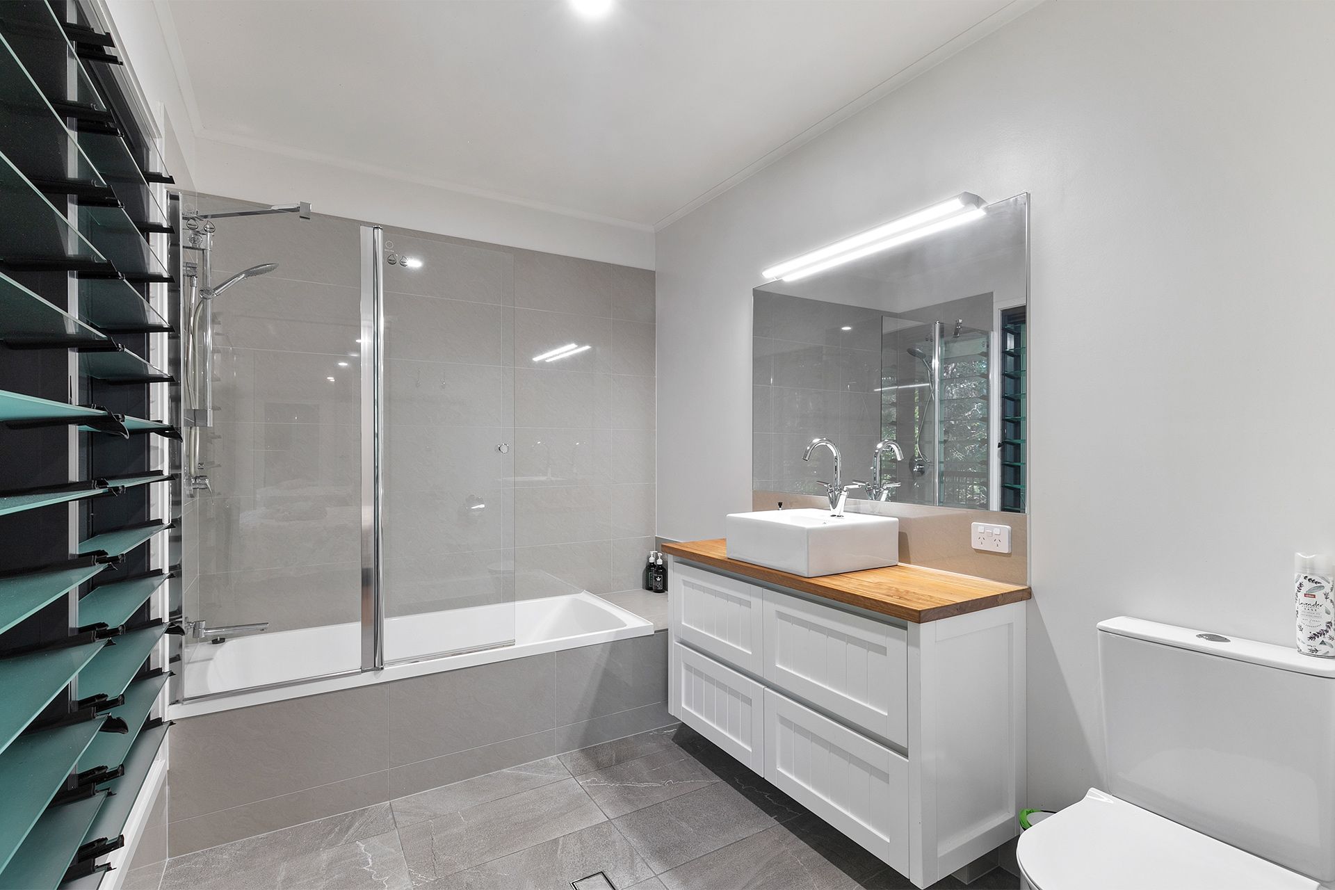 Modern bathroom with white vanity, wooden countertop, and gray tiled walls and floor. Includes a bathtub with glass shower screen, a toilet, and a large mirror.